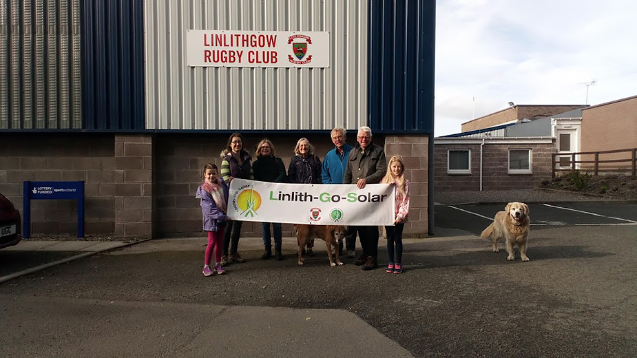 People holding up a sign 'Linlith-Go-Solar' in front of a Ruby Club building and a golden retriever beside them.