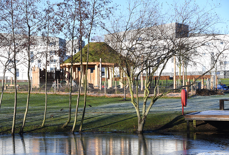 An open circular outdoor building with a grass roof and pond and trees at the forefront.