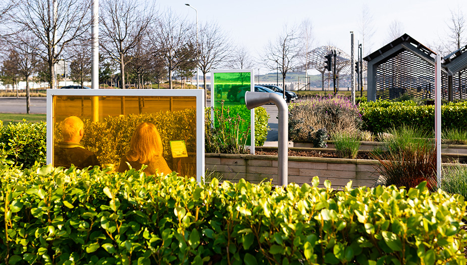 A photograph of two people on a park bench surrounded by wooden planters, art installations and two colourful glass screens 