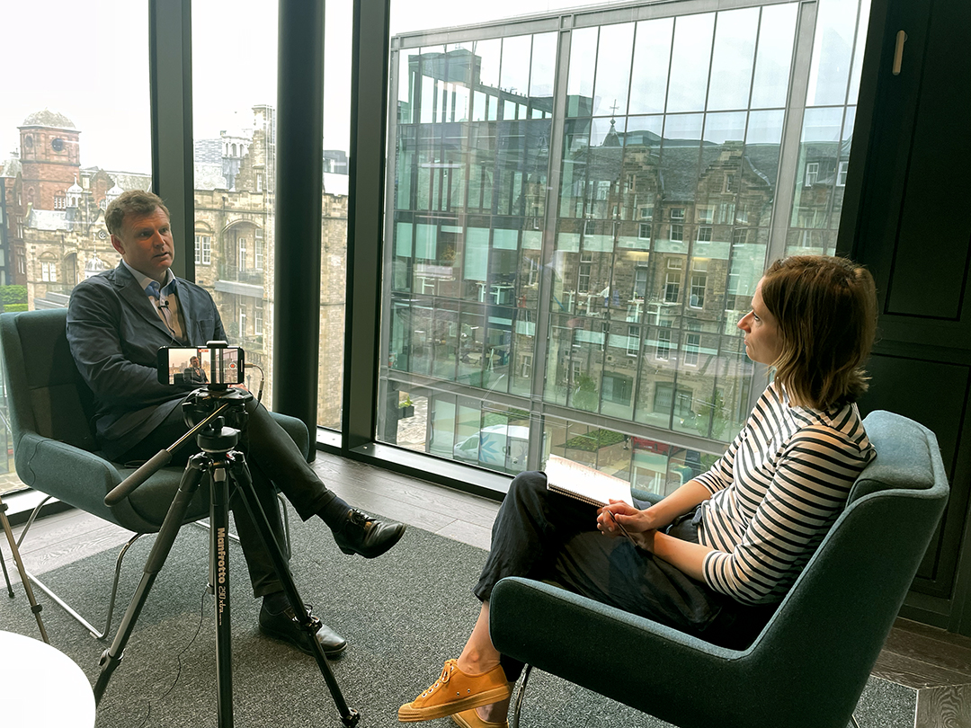 A man wearing a suit sitting in a chair in a glass building, facing a woman in a striped top with a camera phone on a tripod between them. 