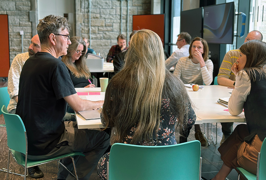 a group of people sitting down around a table working together in a workshop with pens, paper and sticky notes on a table