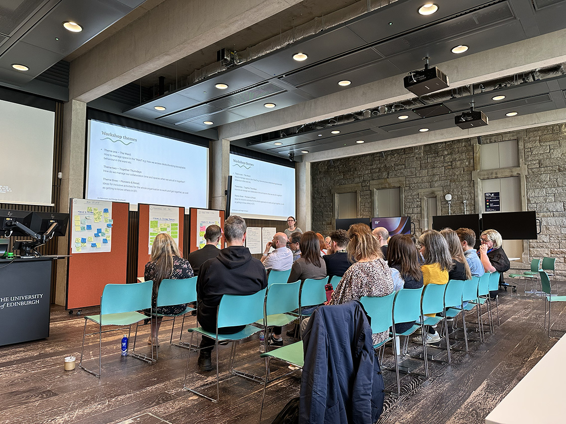 A lecture theatre with the backs of group of people watching a speaker with two large screens above her