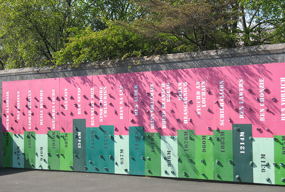 A colourful climbing wall with names and heights of famous Scottish Munros listed vertically on pink and green panels against a stone wall.