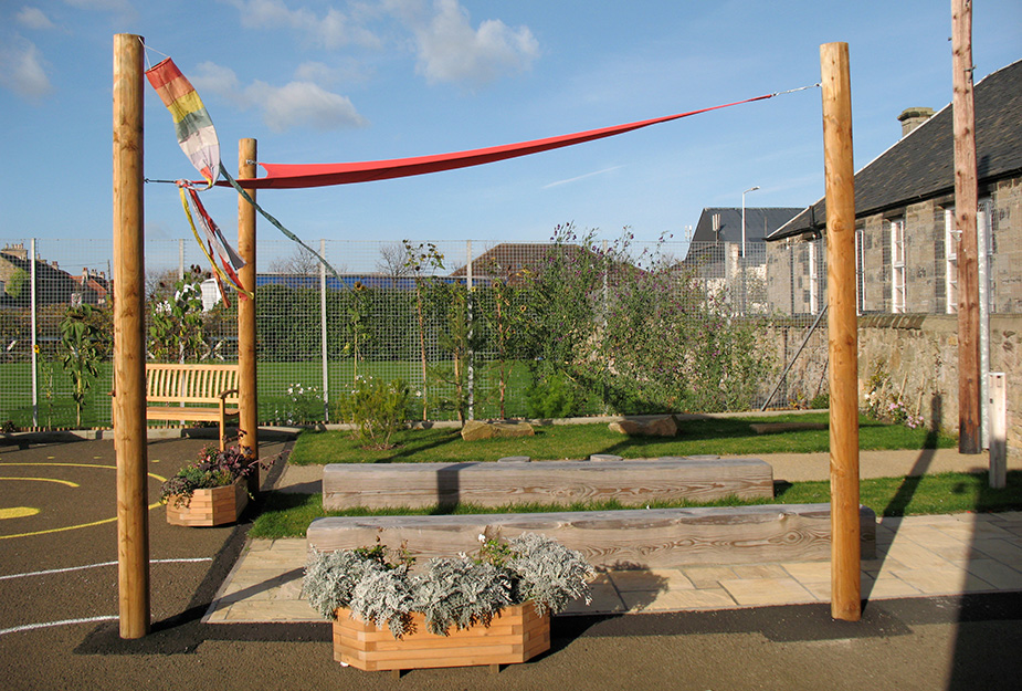 A seating area in a school ground featuring a red canopy, colourful flags, and two parallel timber seating.