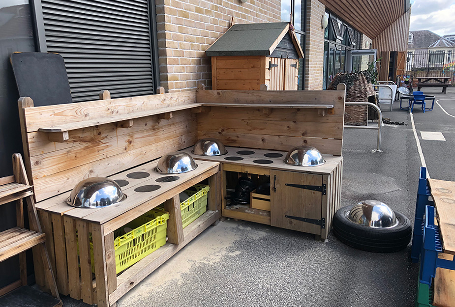 Outdoor play area in a school ground featuring a mud kitchen made out of timber with silver bowls, storage baskets and a small storage unit in the background.