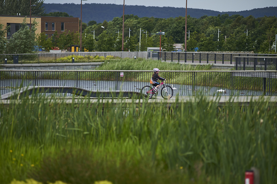 A young girl cycles passed wooden panels springing out of the floor to create fancy benches at Inverness College University of the Highlands and Islands landscape encourages learning activities to ‘spill’ out of the building.