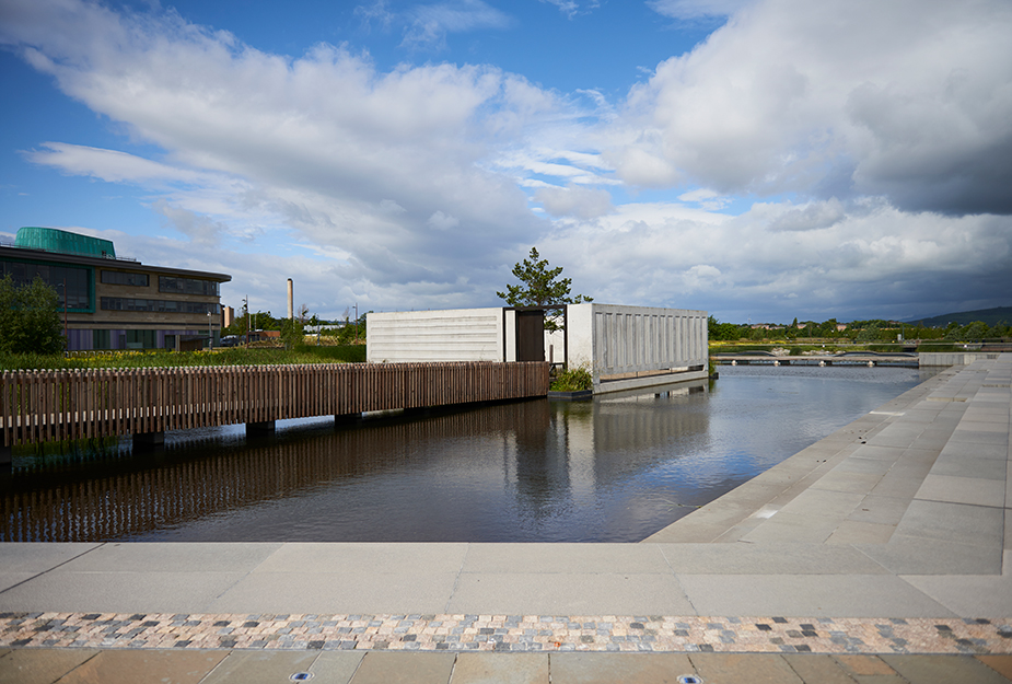 A grey concrete-strutted structure sites along water on an outdoor learning opportunitties on ‘An t-Eilean’, (the Island) at UHI, Elgin.
