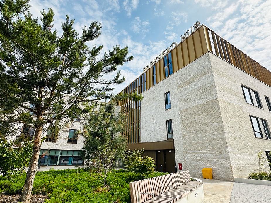 Exterior view of the modern building (Parkhead Hub), surrounded by greenery and a wooden bench, under a partly cloudy sky.