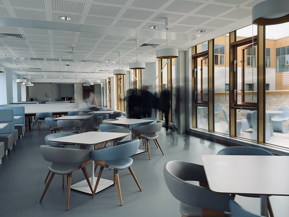 Staff dining interior at Parkhead Hub with minimalistic furniture, bright lighting, and people blurred in motion by the windows.