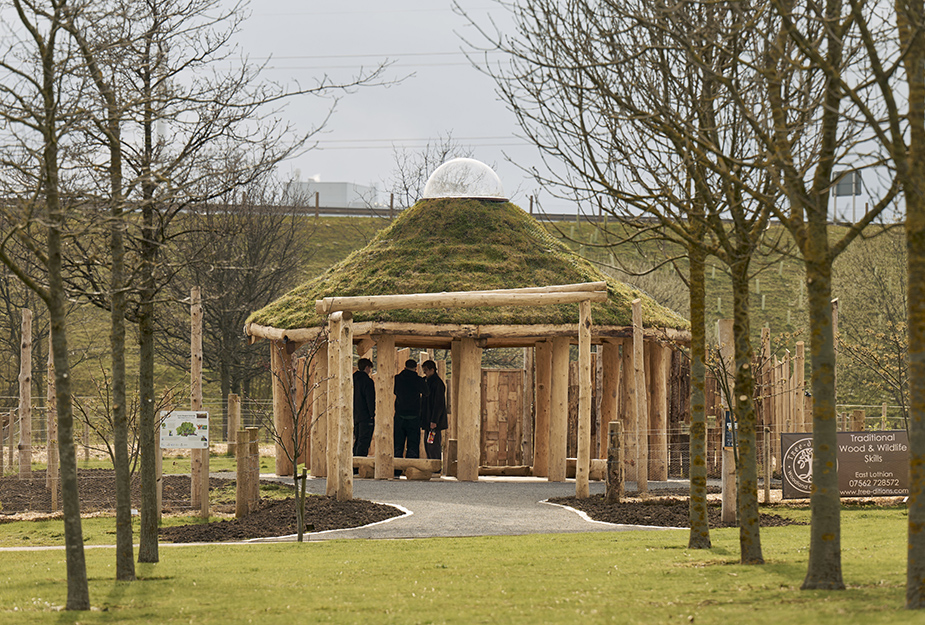 A group of people stands near a wooden structure with a grassy roof, surrounded by trees at the Outdoor Learning Hub at QMU.