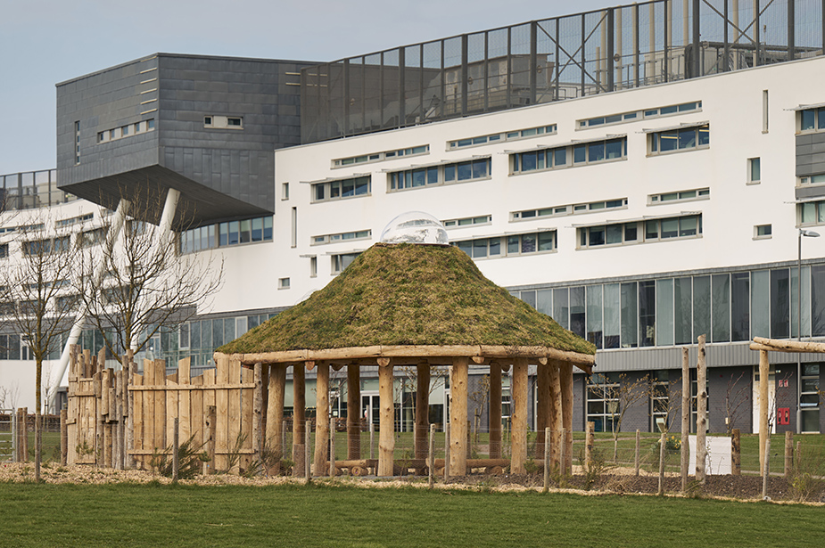 A grassy, dome-shaped pavilion (The Howff) made of wood sits in a green space beside a modern, sleek building university building.