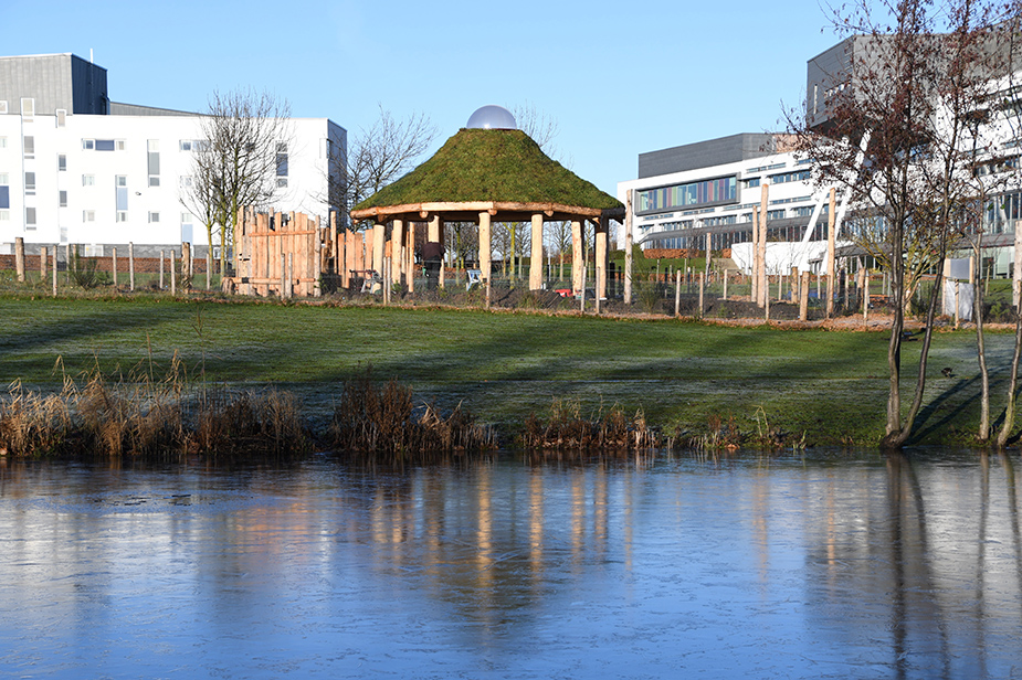The Howff with its living roof, covered in grass near a pond reflecting surrounding buildings and trees during the winter.
