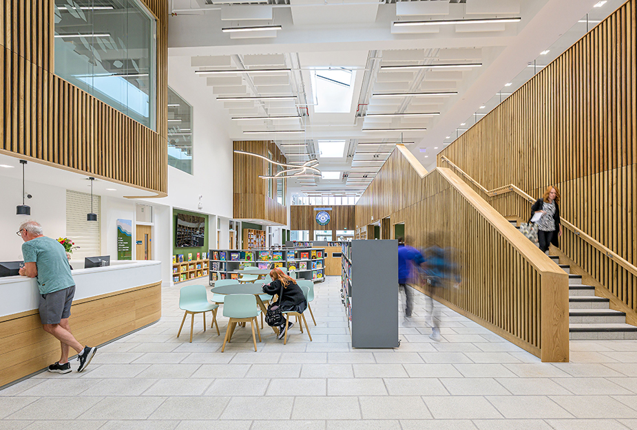 Modern school interior with wooden paneling, white walls, seating area, bookshelves, and people interacting and walking.