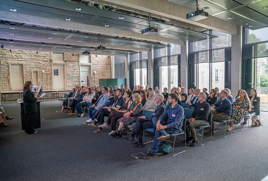 A crowd of people sat down in a modern building listening to a presentation with full length windows in the background.