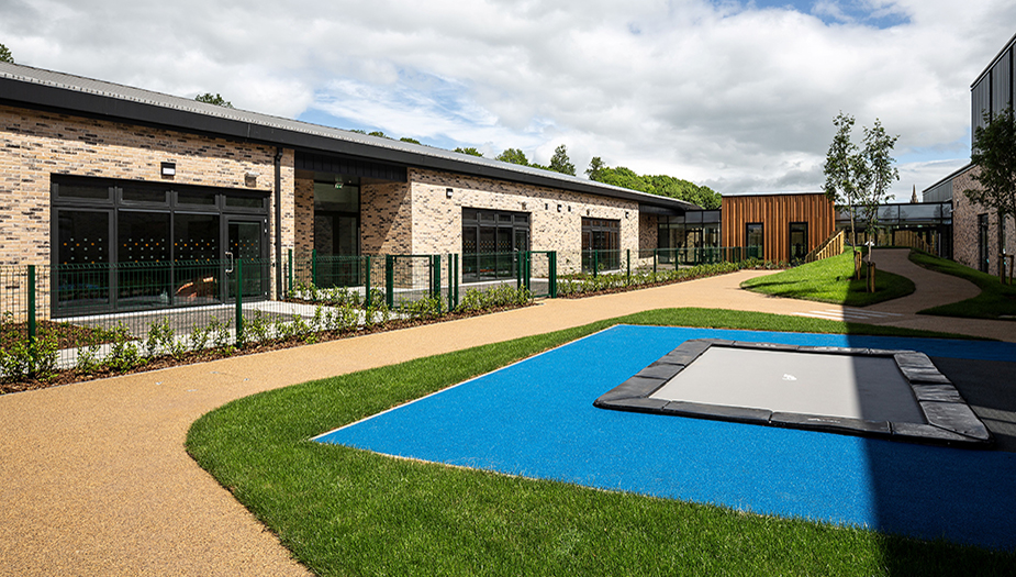 Modern school courtyard with brick buildings, blue trampoline, green grass, paved pathway, and partly cloudy sky.