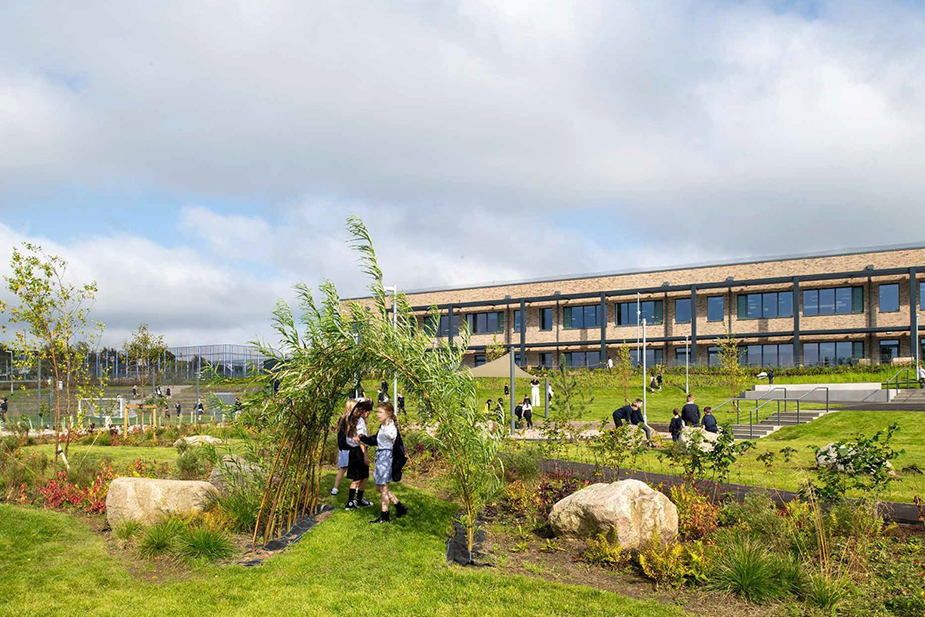 School playground with students walking and sitting near a grass-covered arch and a modern brick school building under cloudy sky.