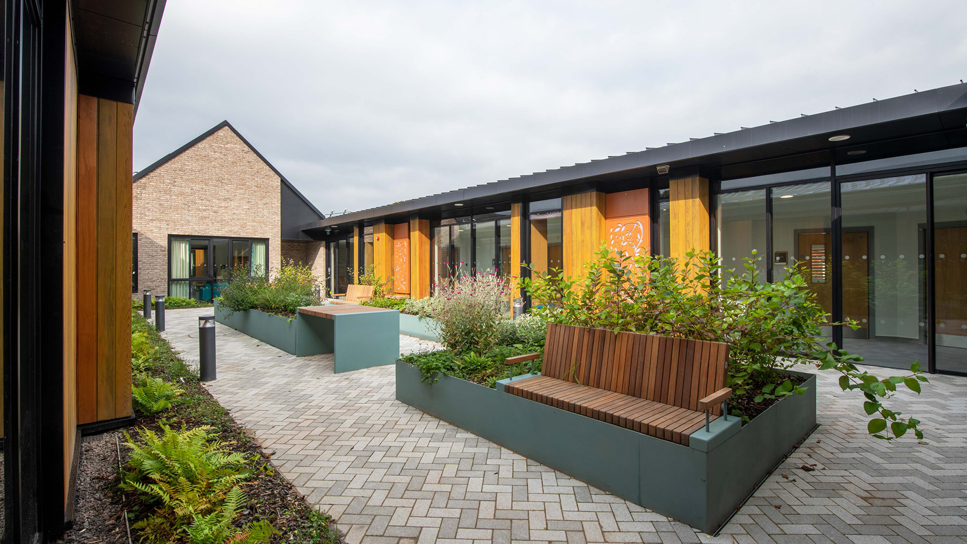 Modern hospital courtyard with wooden benches, raised flower beds, paved walkway, and greenery under a cloudy sky.