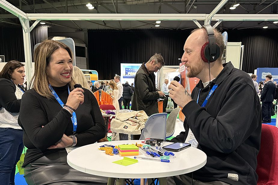 Two people conducting an interview for a podcast at a busy conference or trade show setting.