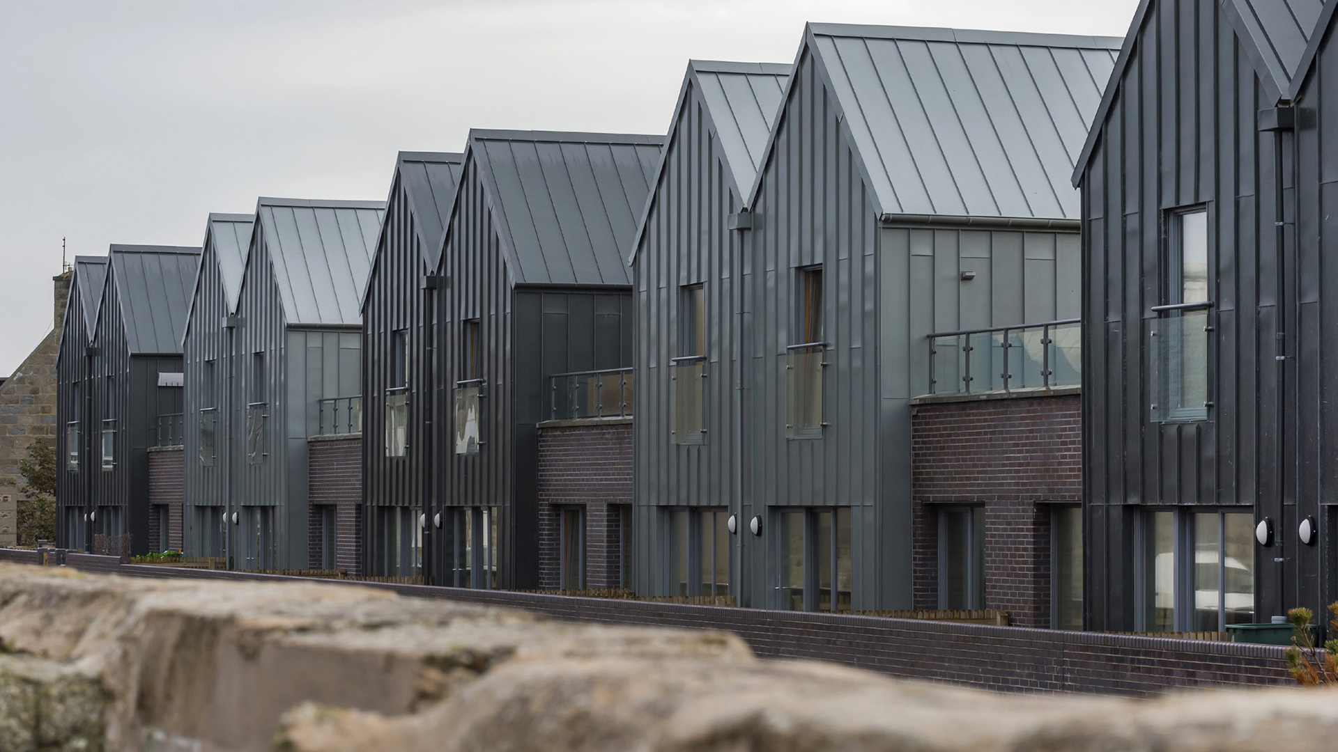 A row of interlocking houses in Fraserburgh with dark and light grey metal cladding and dark brick exterior walls beside each unit. 