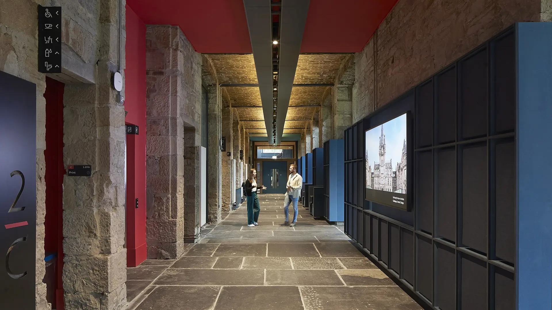 University hallway with exposed stone walls, red ceiling accents, exhibit screens, and two people walking through the hallway.