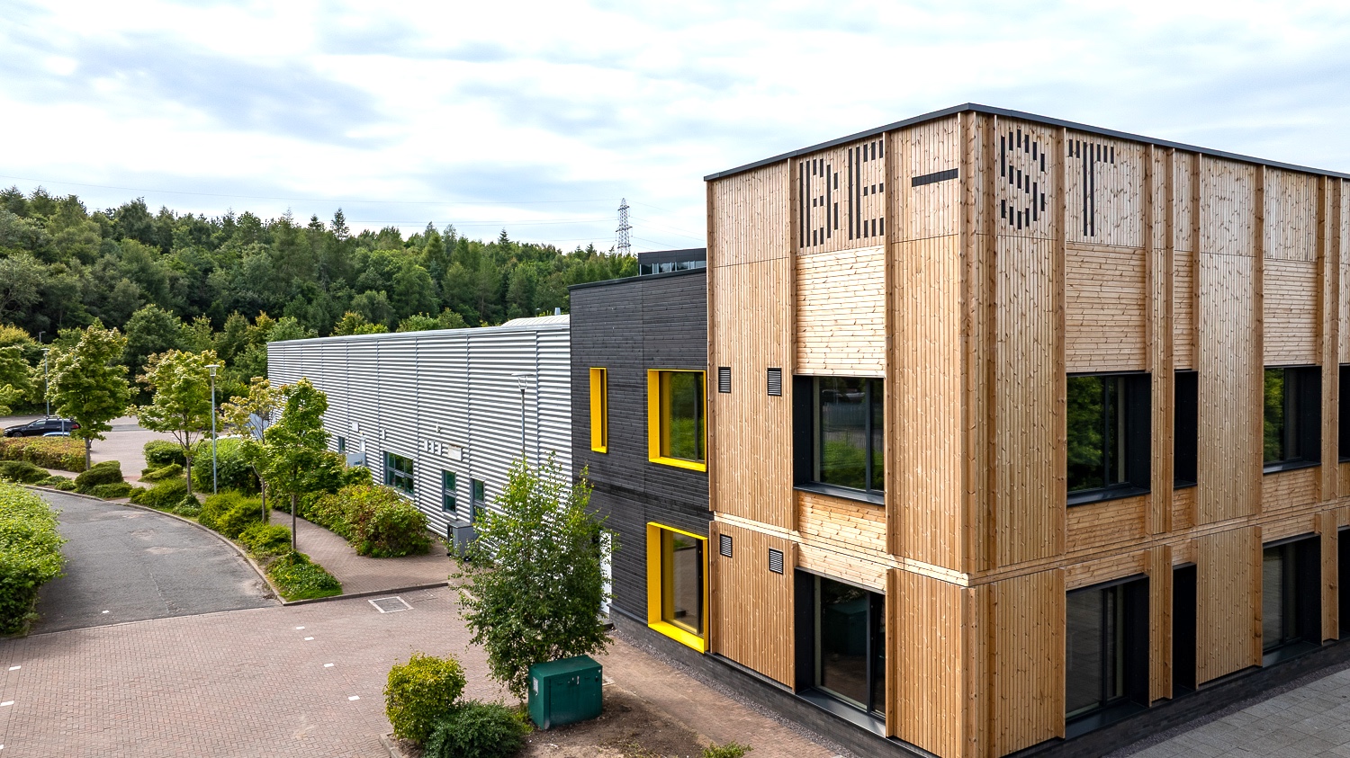 Modern wood-clad and metal commercial buildings with "BE-ST" painted onto the wooden cladding, beside landscaped walkways and a wooded hillside.