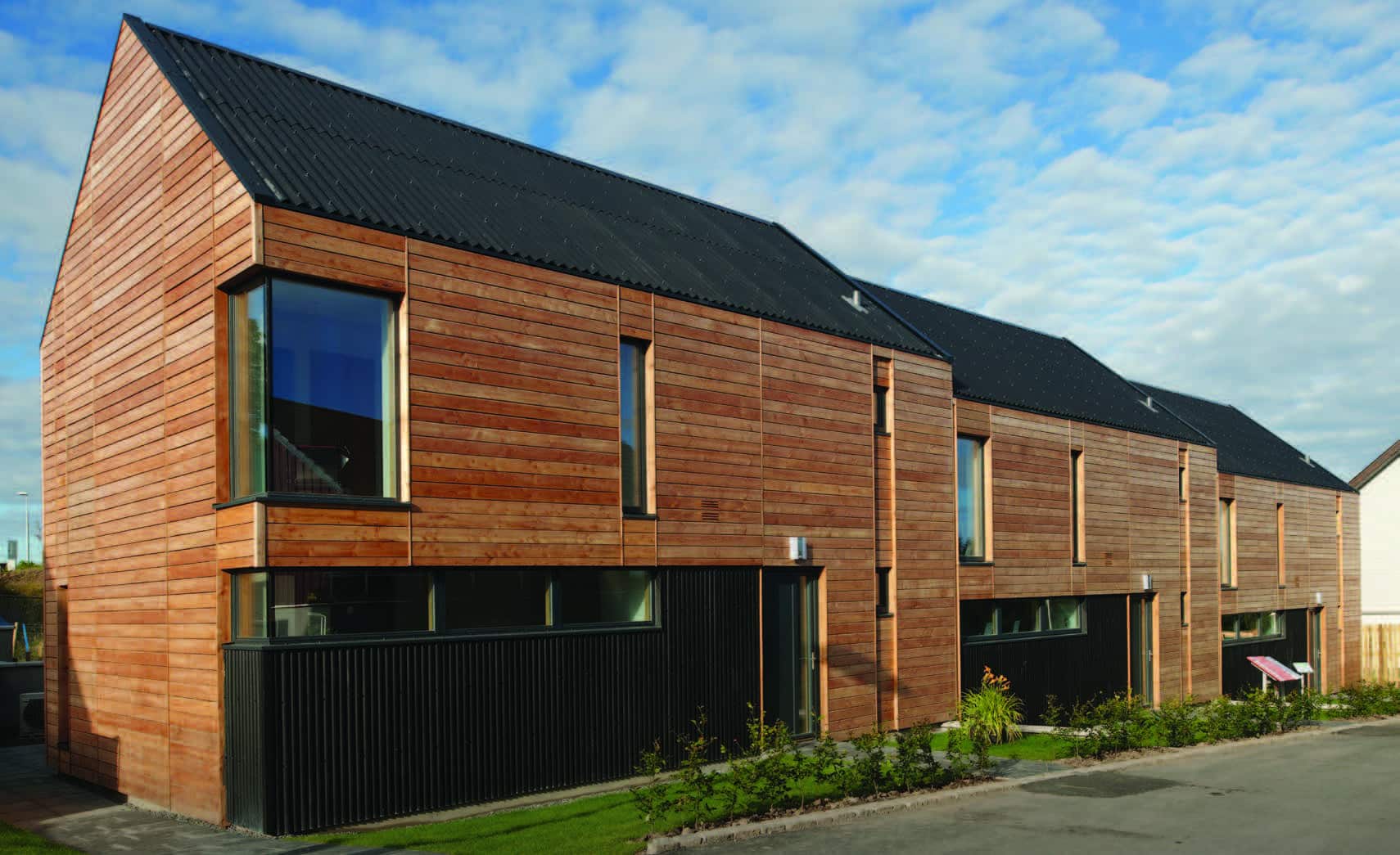 Modern row of wood-clad PassiveHaus homes with black roofs and large windows under a partly cloudy blue sky.