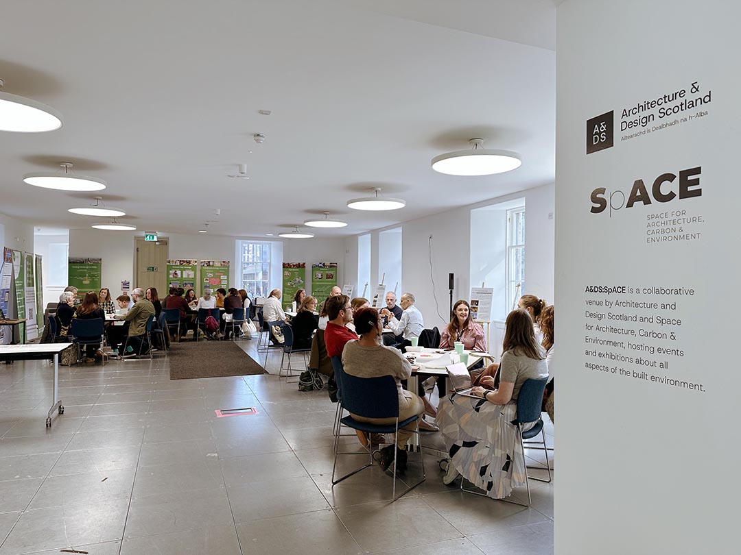 People sat around tables in groups at an event with the Architecture and Design Scotland and SpACE logo in the foreground.